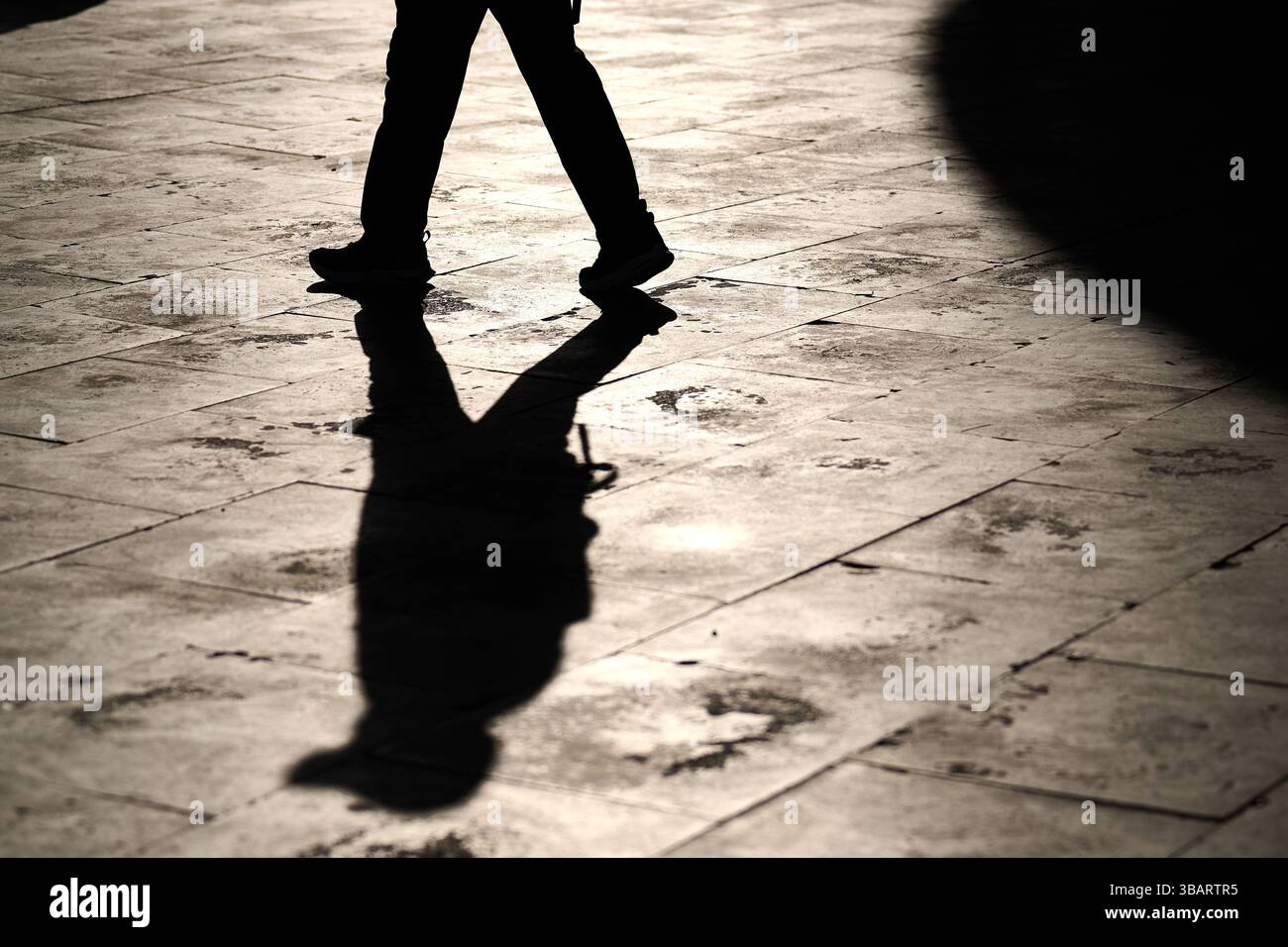 Una persona che cammina da sola su piastrelle illuminate dal sole con una silhouette affilata e un'ombra allungata che si estende sul terreno. Foto Stock