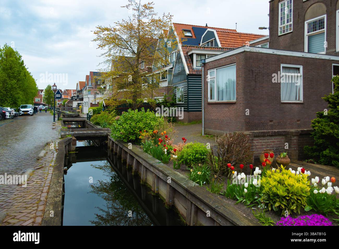 Colorato canale di Volendam, Paesi Bassi, con edifici tradizionali, fiori in fiore, alberi, e un piccolo ponte sotto un cielo nuvoloso. Foto Stock