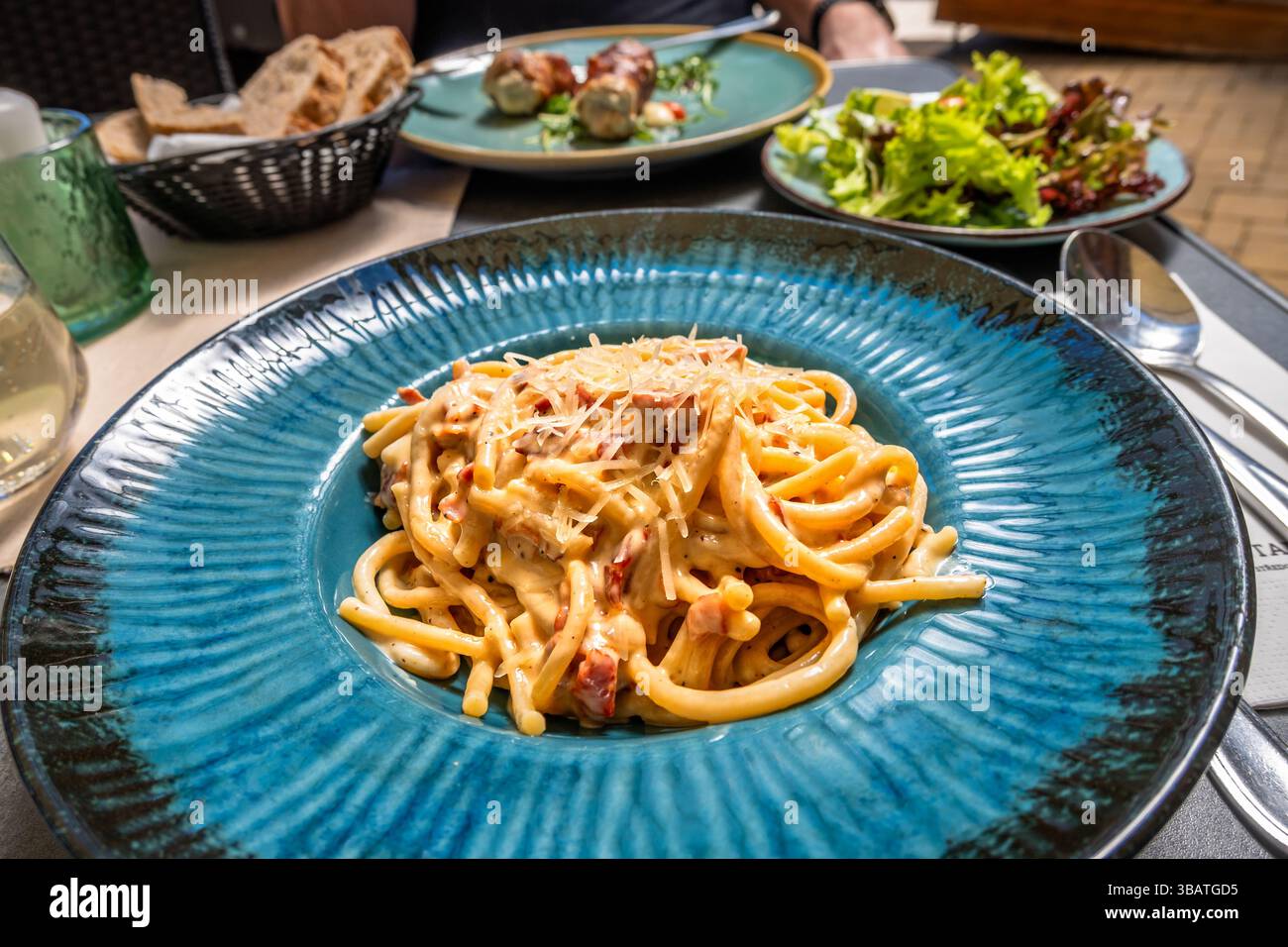 Spaghetti alla carbonara in piatto blu, insalata, pane, bevanda e involtino di maiale sul secondo piatto sul tavolo del ristorante. Foto Stock