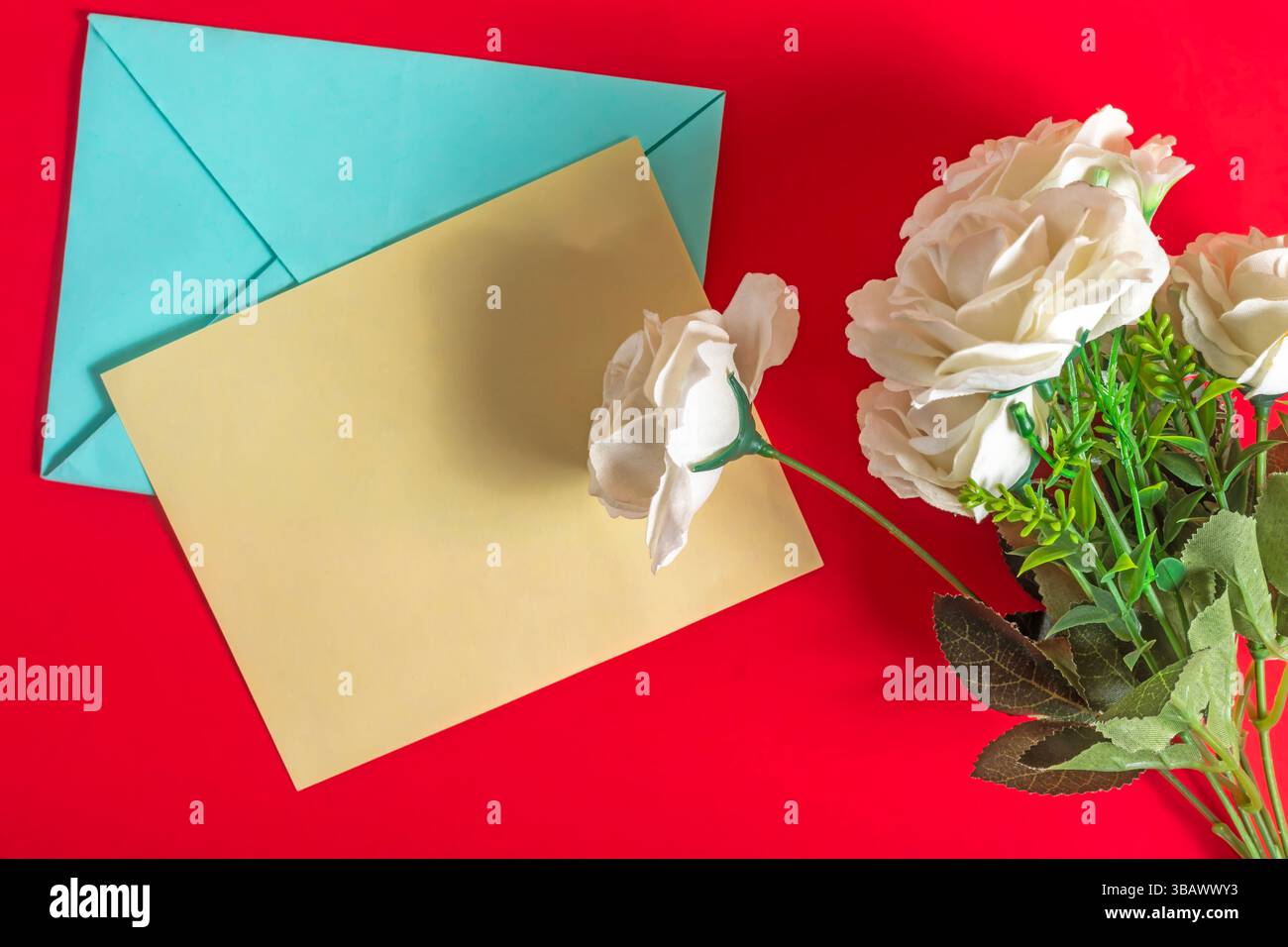 Una disposizione di carta, busta e rose bianche su un'accattivante superficie rossa, perfetta per feste, messaggi romantici o serate ponderate Foto Stock
