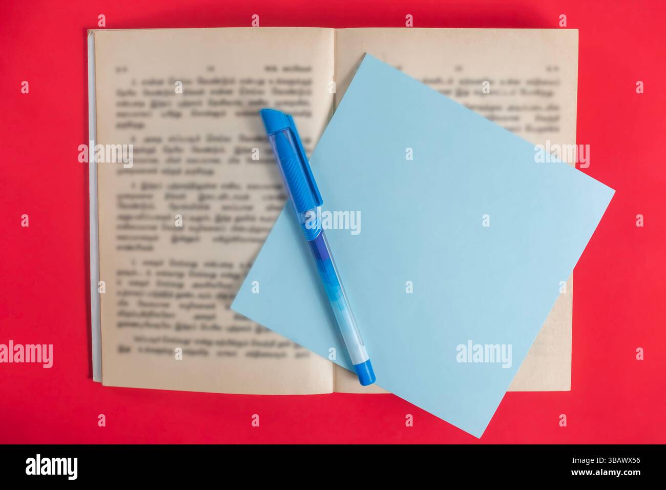 Vista dall'alto di un libro aperto con testo sfocato, nota adesiva blu e penna su un vivace sfondo rosso che rappresenta l'istruzione, la conoscenza e lo scopo di scrittura Foto Stock