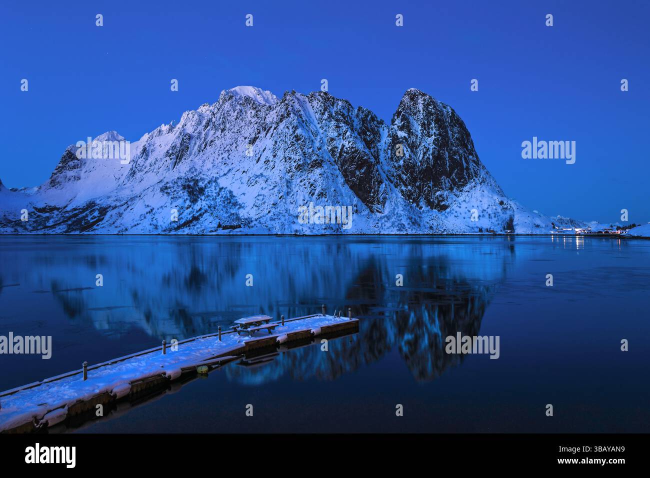 La vetta della montagna Olstind sopra il Kirkefjord nell'ora blu con una tinta di tramonto, un molo di legno in primo piano, Reine, Isole Lofoten, Norvegia Foto Stock
