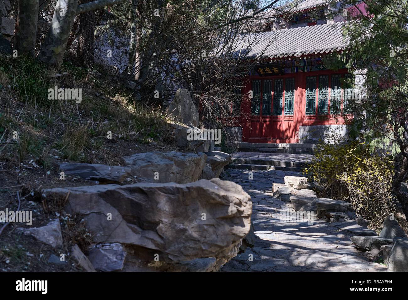 Un tranquillo sentiero in pietra conduce attraverso una foresta ombreggiata a un vecchio edificio cinese dalle mura rosse nascosto tra alberi e rocce Foto Stock