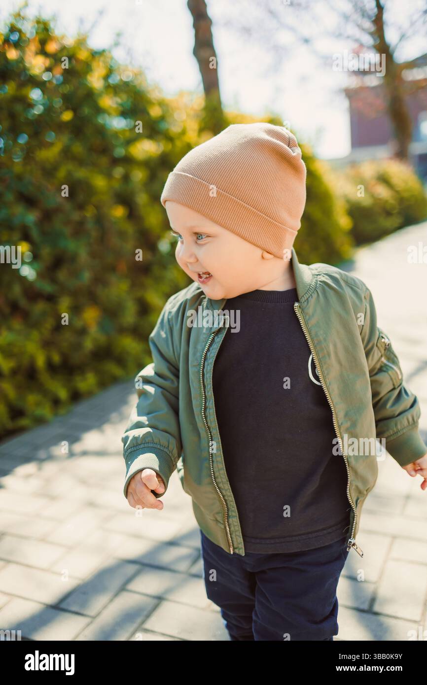 Un bambino allegro sta godendo una giornata di sole su un sentiero, indossando un bomber alla moda e un cappello accogliente, circondato da una vibrante vegetazione che esalta la a Foto Stock