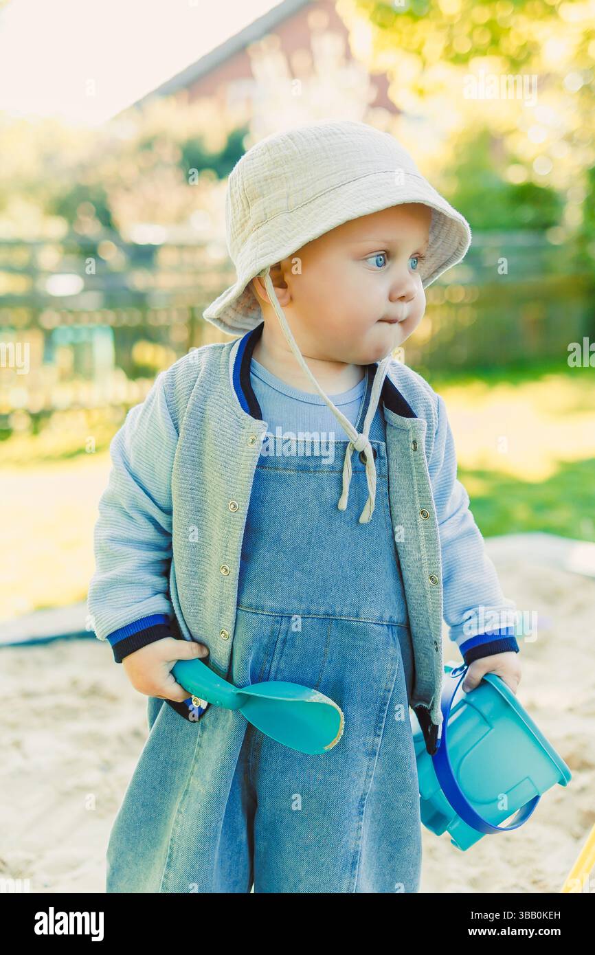 Un bambino allegro vestito con un intimo vestito esplora felicemente una sandbox in una giornata di sole, circondato da giocattoli colorati e da una vivace fille sul retro Foto Stock