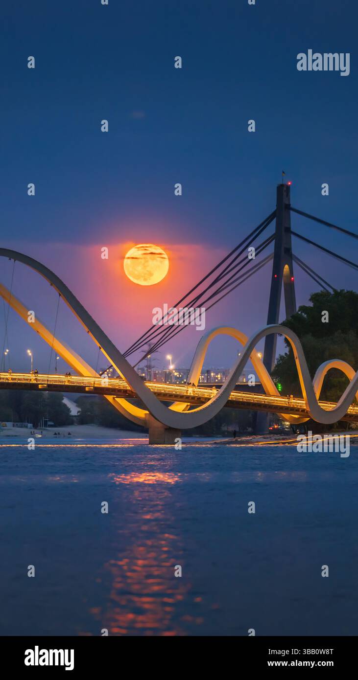 La luna piena di fiori si innalza sul ponte pedonale di Kiev, Ucraina, gettando una calda luce arancione sul fiume Dnipro in una tranquilla notte di maggio Foto Stock