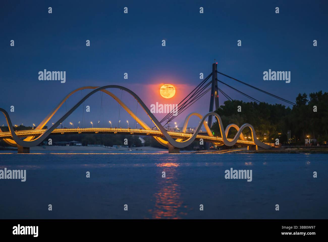 La luna piena di fiori si innalza sul ponte pedonale di Kiev, Ucraina, gettando una calda luce arancione sul fiume Dnipro in una tranquilla notte di maggio Foto Stock