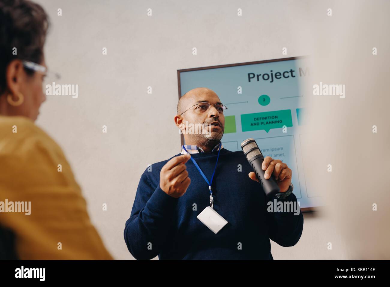 Un uomo d'affari professionista discute l'ambito di un progetto con i colleghi in un ambiente formale, utilizzando un microfono per chiarezza. La riunione presenta una visua Foto Stock