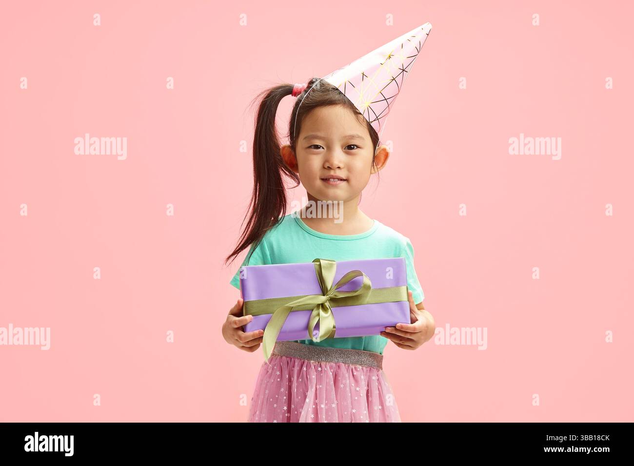 Affascinante ragazza asiatica per bambini di circa 5-6 anni in cappello da festa, indossa abiti festivi, regge la confezione regalo su sfondo rosa isolato Foto Stock