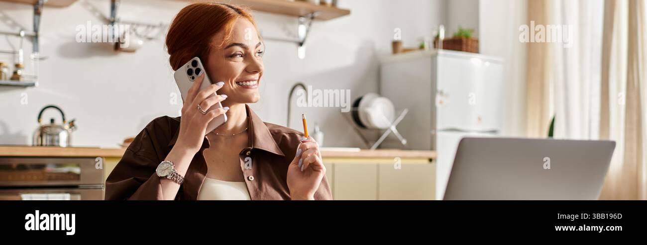 In una cucina luminosa, una vivace donna dai capelli rossi chatta al telefono, sorridendo e tenendo in mano una penna. Foto Stock