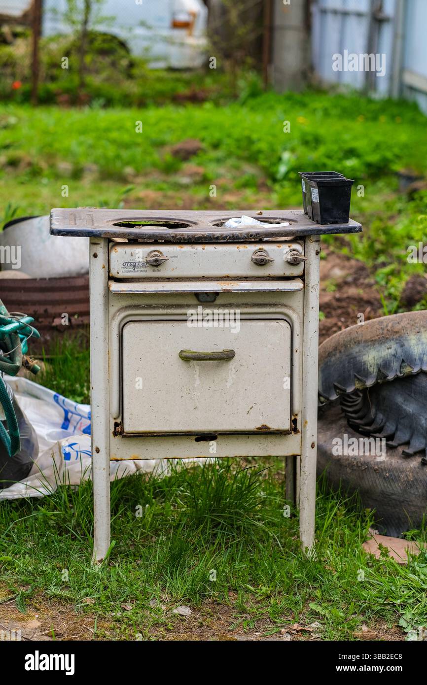 mosca, russia, 10.05.2025 Una vecchia stufa arrugginita si trova in un cortile erboso. La stufa è circondata da vari oggetti, tra cui uno pneumatico, una pianta in vaso e un Foto Stock