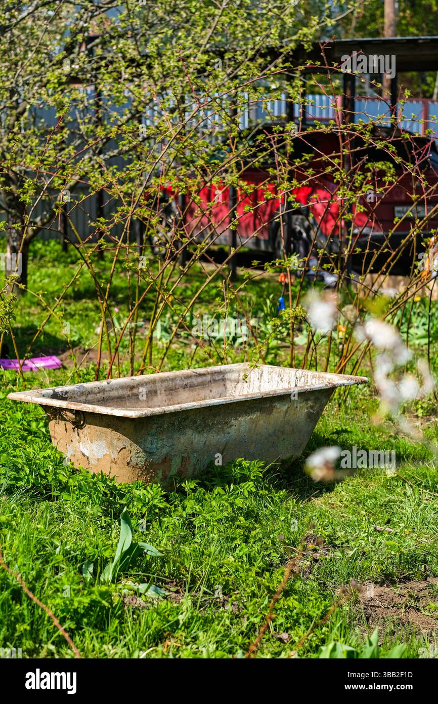 mosca, russia, 10.05.2025 Una grande e vecchia vasca da bagno si trova in un cortile erboso. Il cortile è pieno di fiori e cespugli, e c'è un camion rosso nel retro Foto Stock