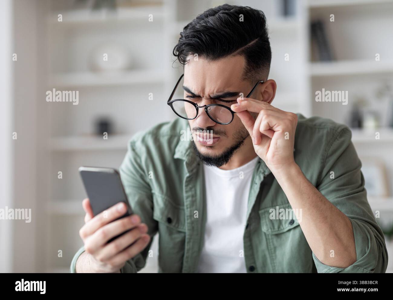 Problemi di vista concetto. Giovane uomo arabo in occhiali guardando lo schermo dello smartphone Foto Stock