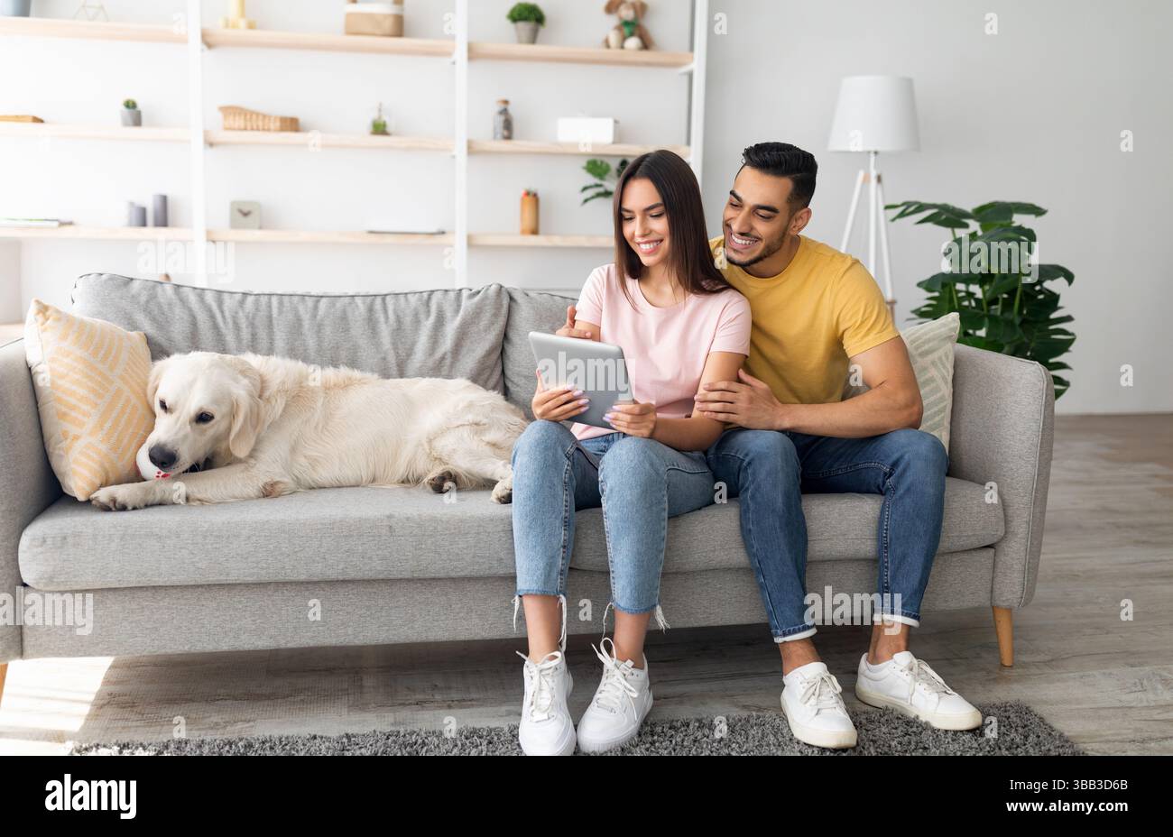 Bella giovane e variegata coppia seduta sul divano con il suo simpatico cane, che usa un tablet pc e naviga sul Web a casa Foto Stock