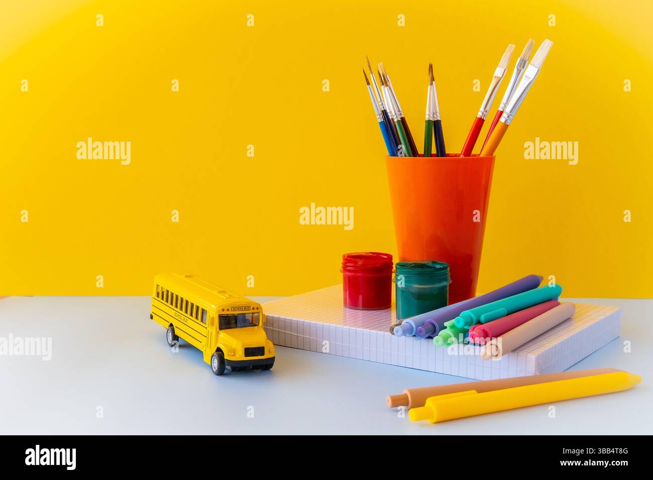 Forniture creative per la scuola disposizione con pennelli penne colorate scuolabus e quaderni su sfondo giallo che rappresentano l'apprendimento artistico Foto Stock