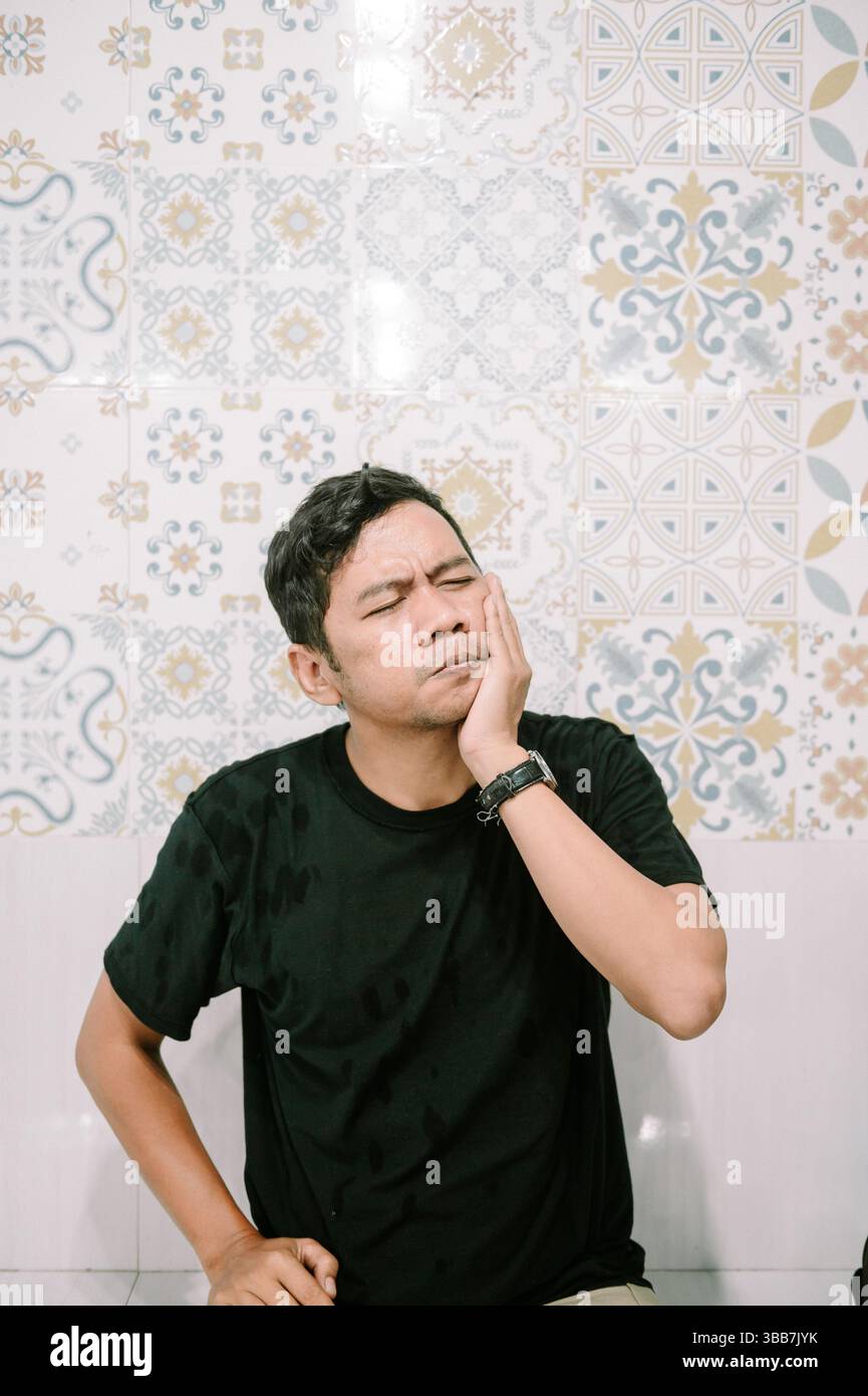 Un uomo malato che tiene il viso con un'espressione dolorosa a causa di un mal di denti su uno sfondo piastrellato ornato Foto Stock