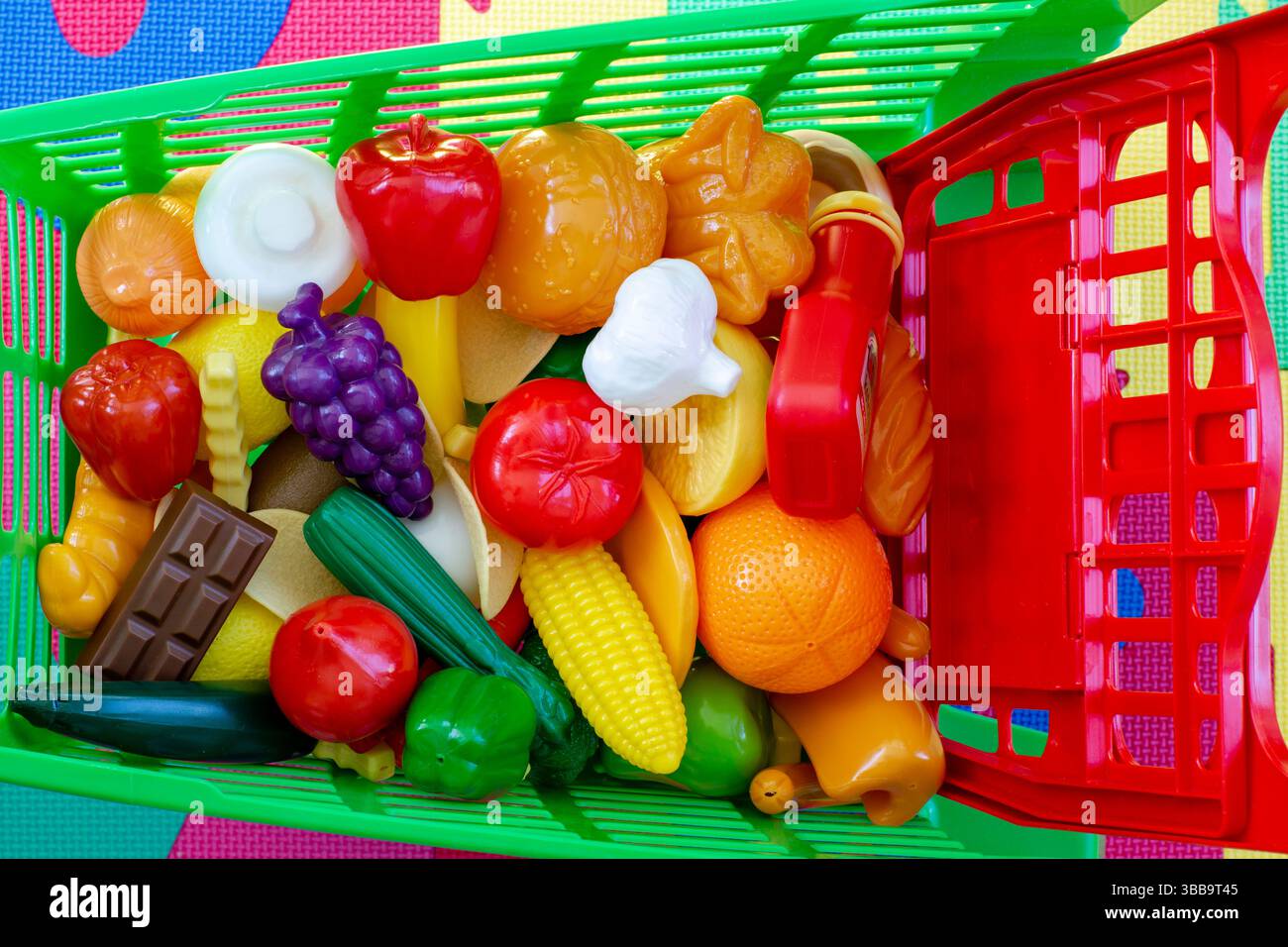 Un vivace cestino per la spesa pieno di coloratissimi cibi da gioco in plastica, tra cui frutta, verdura e spuntini, progettato per bambini fantasiosi Foto Stock