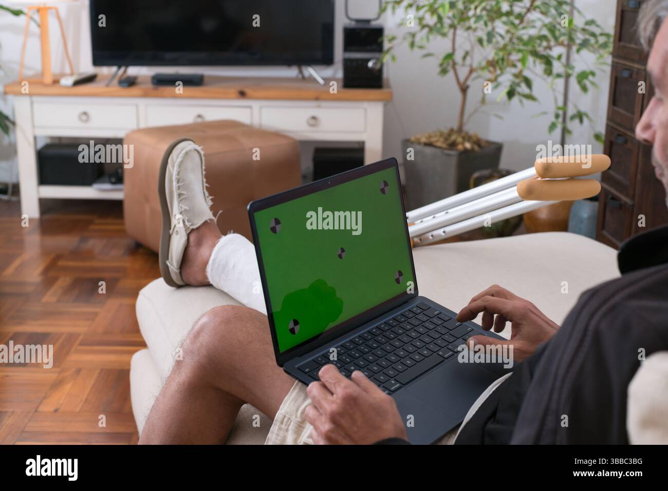 Uomo anziano con gamba rotta e stampelle che utilizza un computer portatile con schermo verde mentre è seduto sul divano di casa Foto Stock