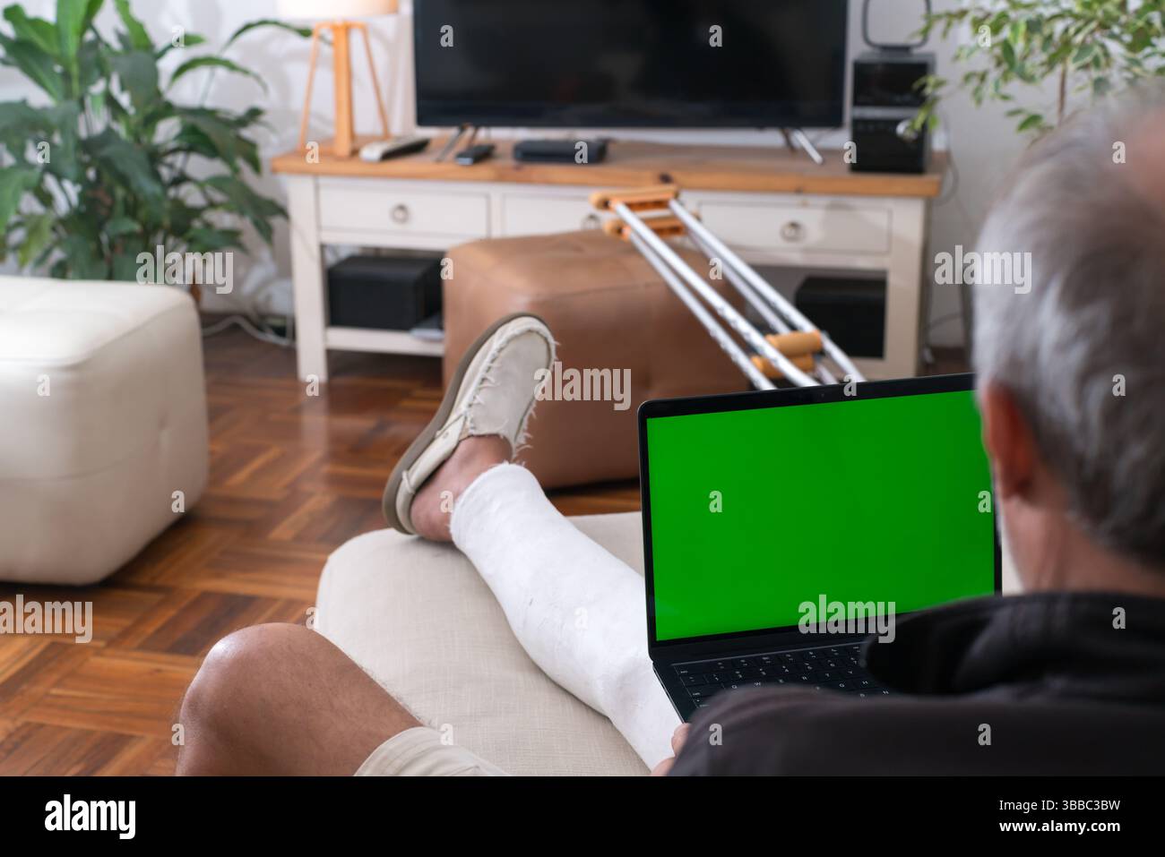 Uomo anziano con una gamba rotta che utilizza un computer portatile con chiave cromatica a schermo verde che si rilassa sul divano di casa Foto Stock