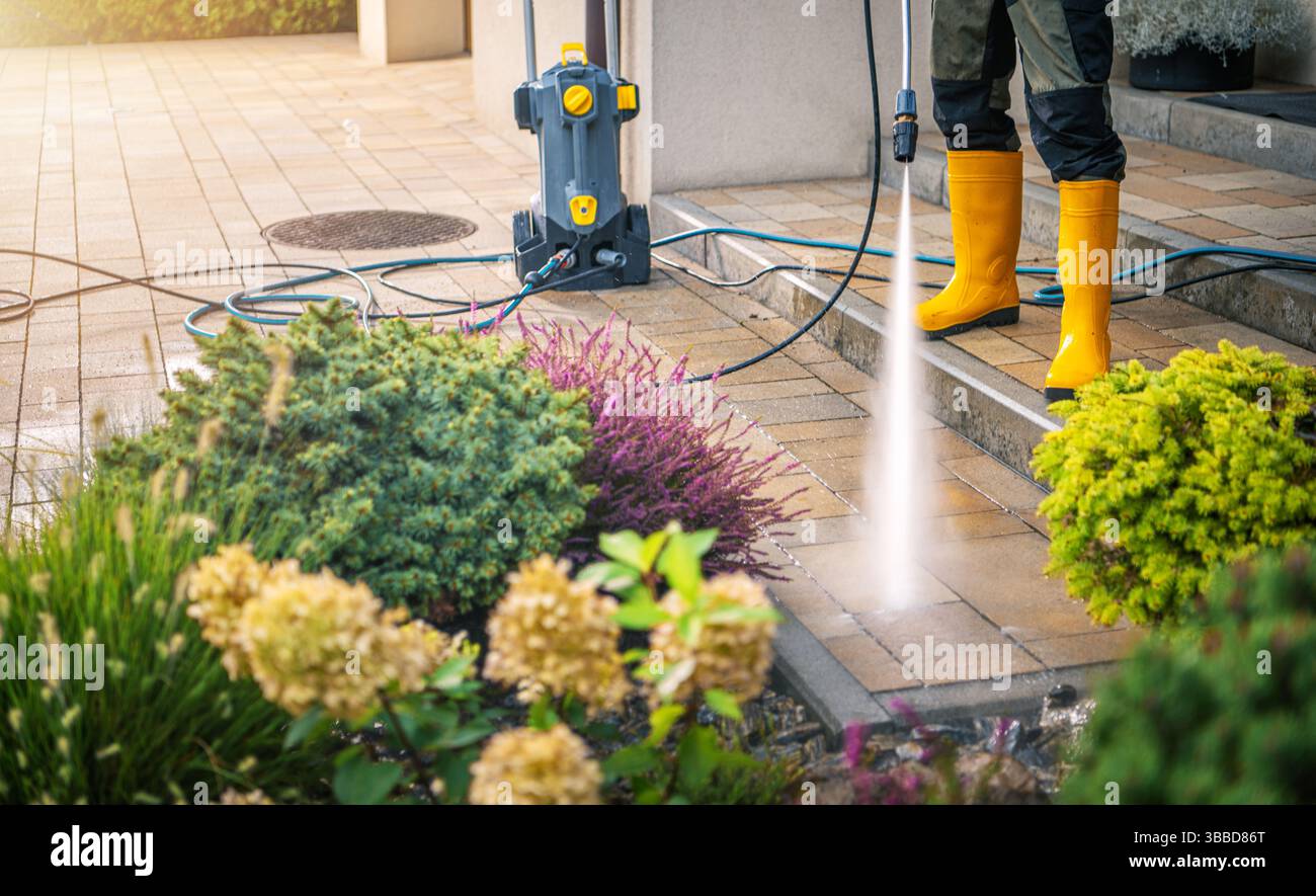 La luce del sole illumina un patio ben tenuto mentre una persona con stivali in gomma gialla utilizza una pulitrice a pressione per pulire la superficie in pietra. Lussureggiante più verde Foto Stock