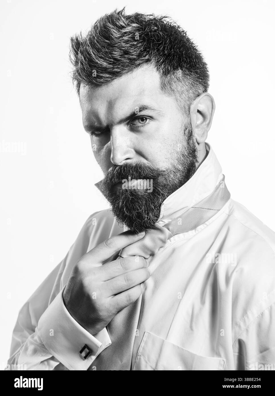 L'uomo elegante con barba e baffi in camicia regola la cravatta sul collo. Sposo in preparazione per il matrimonio. Un uomo d'affari barbuto che si lega alla cravatta. Un uomo d'affari serio Foto Stock