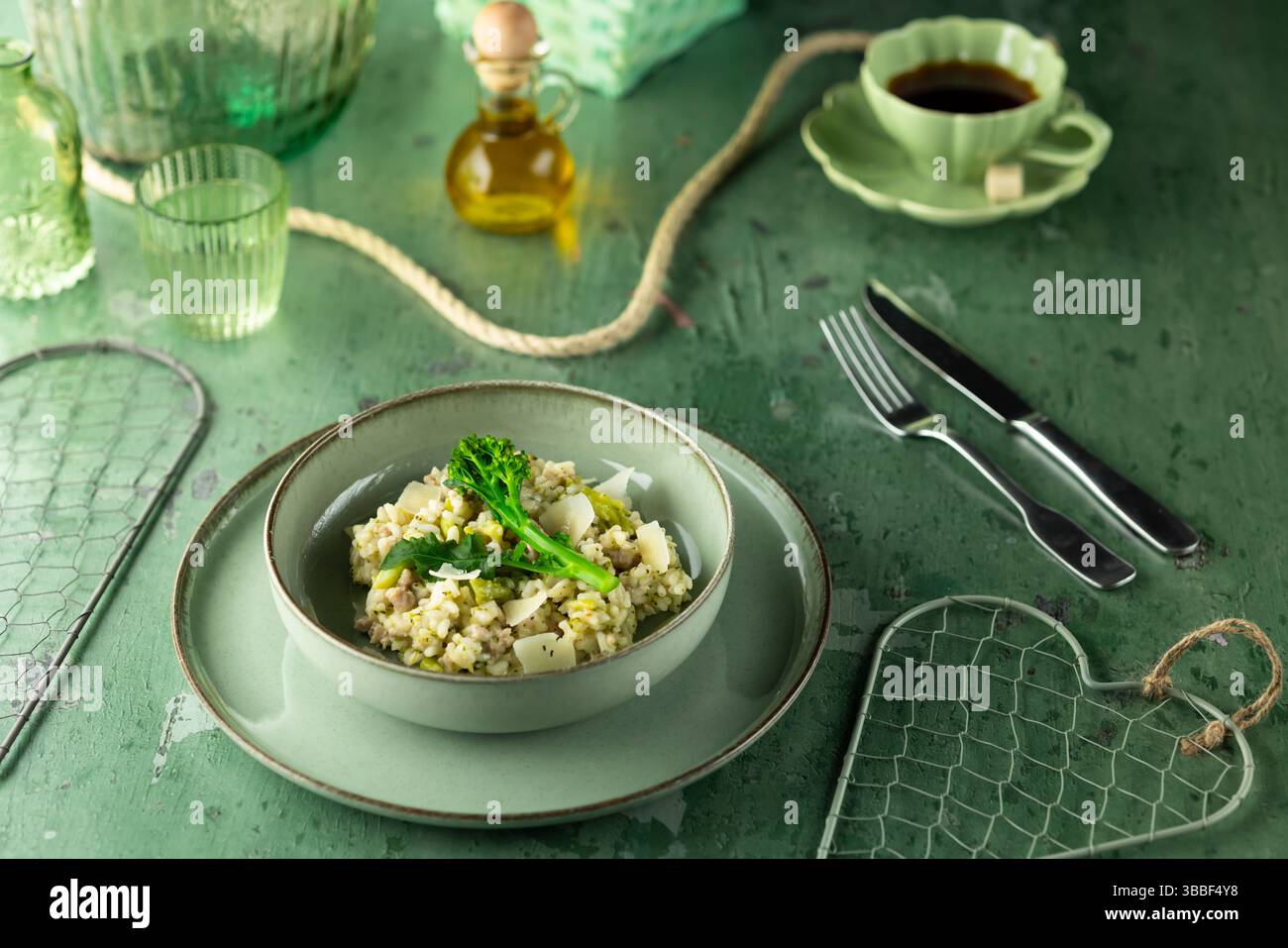 Risotto con broccoli, parmigiano e piselli serviti su piatto verde con forchetta e coltello a parte, olio d'oliva, caffè e tappetini verdi da tavola Foto Stock
