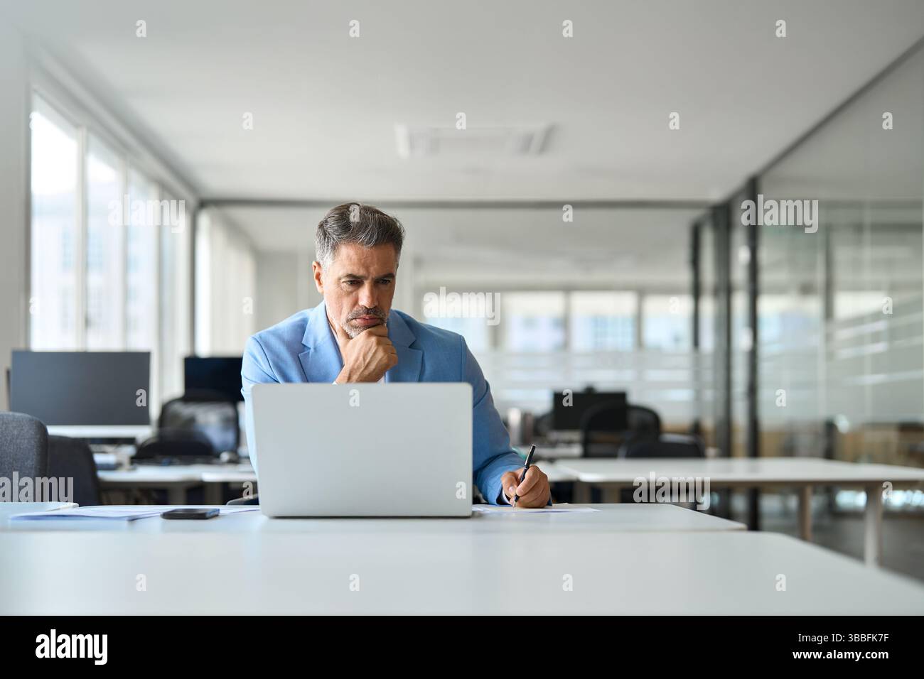 Un uomo d'affari di mezza età, serio e impegnato, che lavora su un notebook e scrive appunti in ufficio. Foto Stock