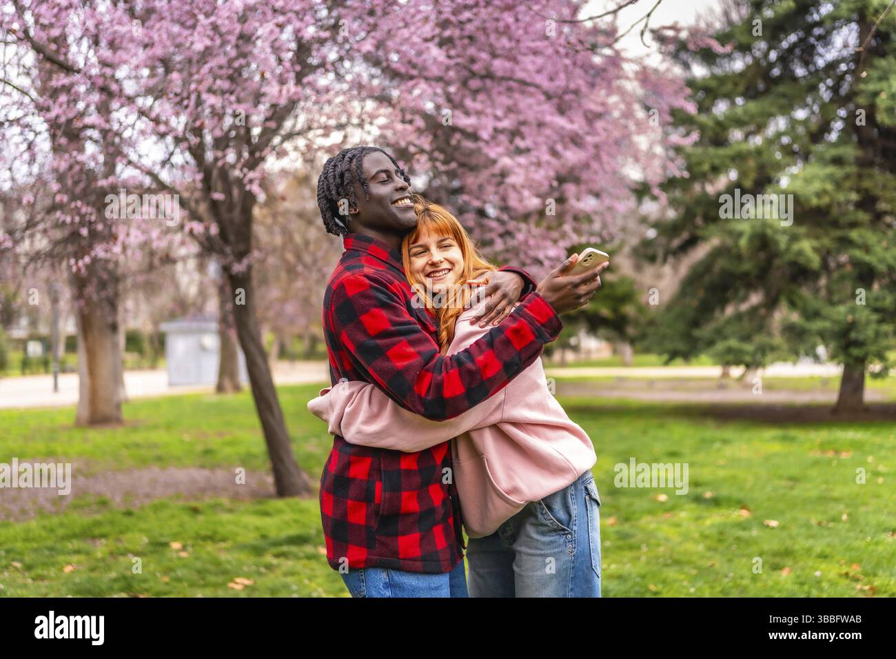 Felice coppia multietnica che scatta un selfie con lo smartphone mentre abbraccia in un parco pieno di alberi in fiore durante la primavera Foto Stock