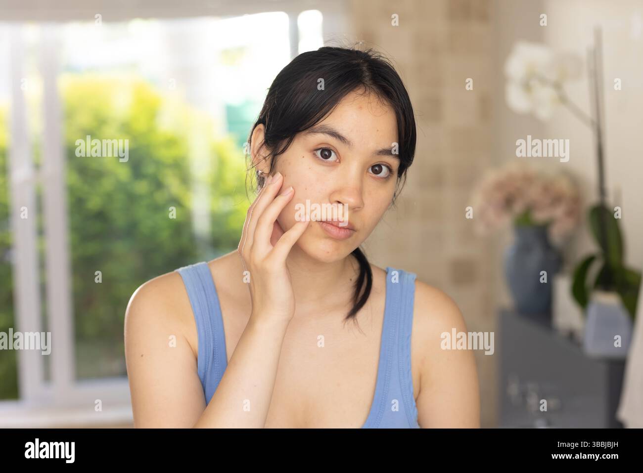 Toccando la guancia destra donna asiatica in piedi in un'area soggiorno accanto alla finestra, con fiori pastello e orchidea Foto Stock