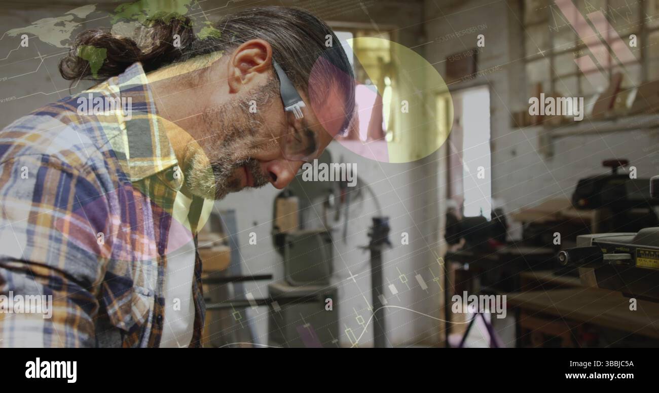 Lavoratore legnaio maturo inclinato che esamina la tavola di legno sul banco di lavoro in un'officina di legno, con occhiali di sicurezza Foto Stock