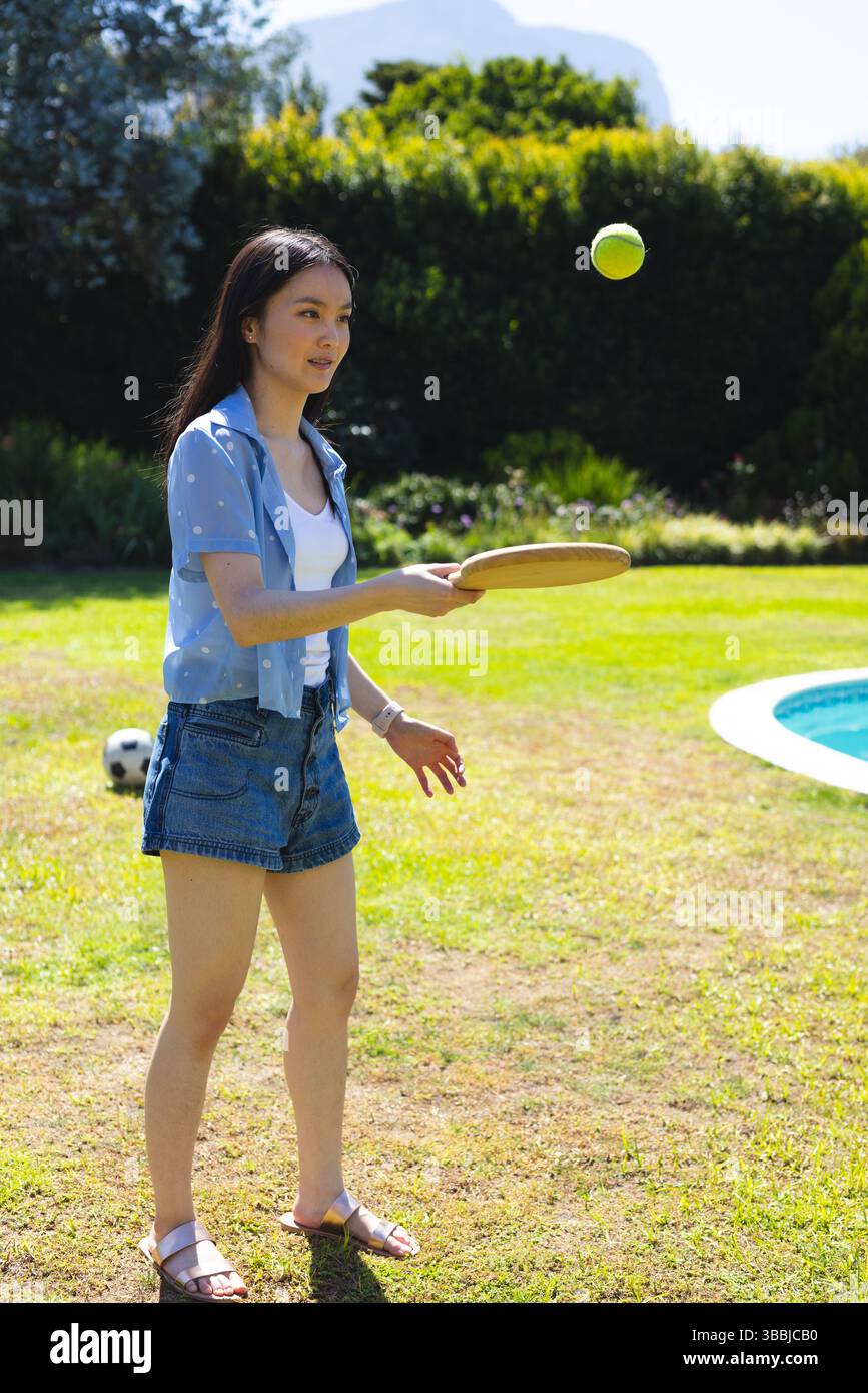 Donna asiatica che rimbalza palla da tennis nel cortile accanto alla piscina a forma di rene, con paddle e pallone da calcio Foto Stock
