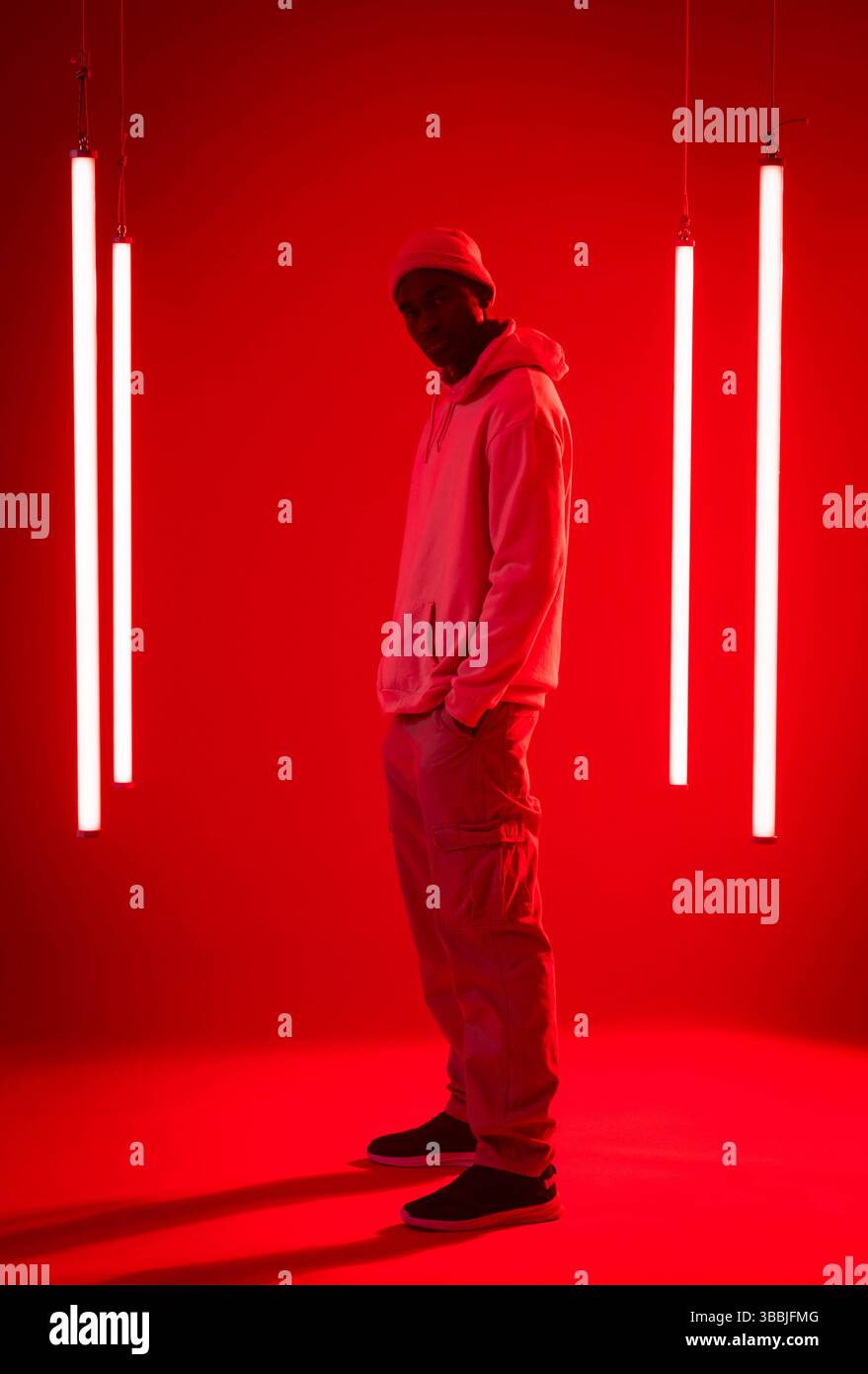 Uomo afroamericano in piedi con cappuccio e scarpe da ginnastica, in posa in studio rosso con luci a tubo Foto Stock