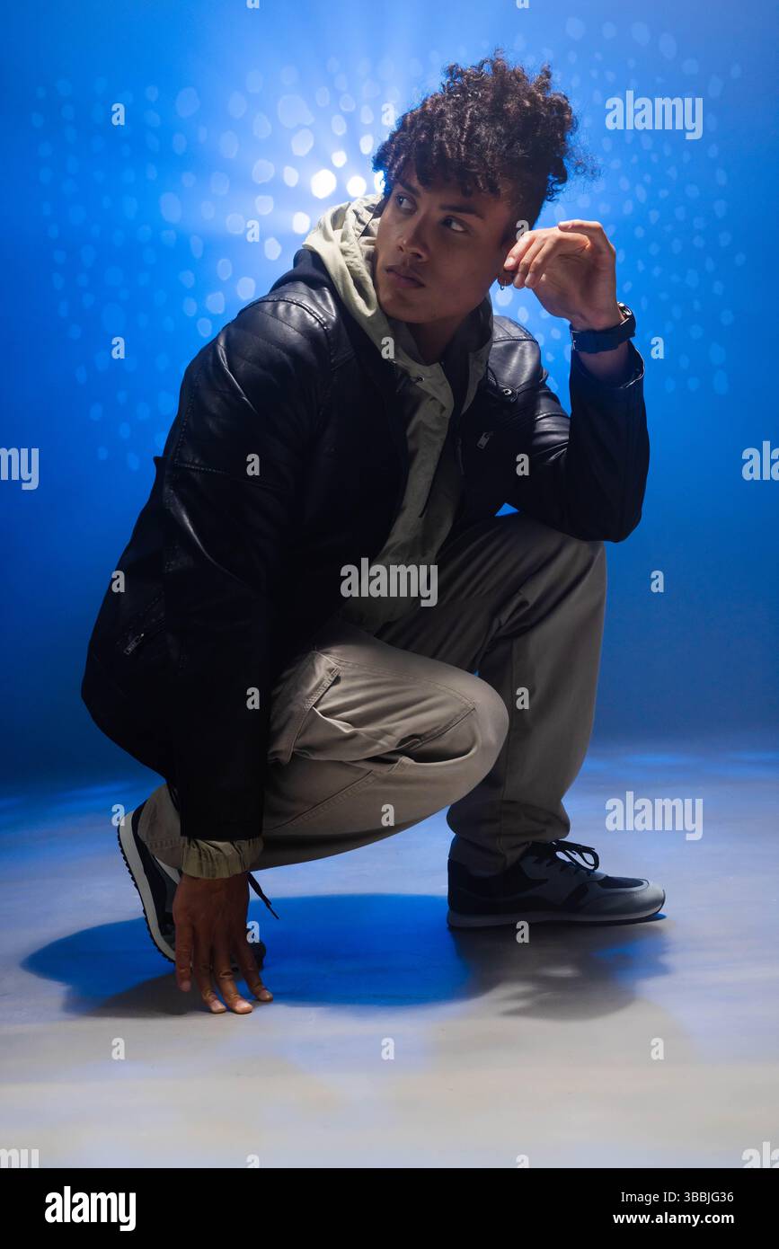 Uomo afroamericano accovacciato che posa in uno studio fotografico, indossa giacca di pelle e pantaloni cargo Foto Stock
