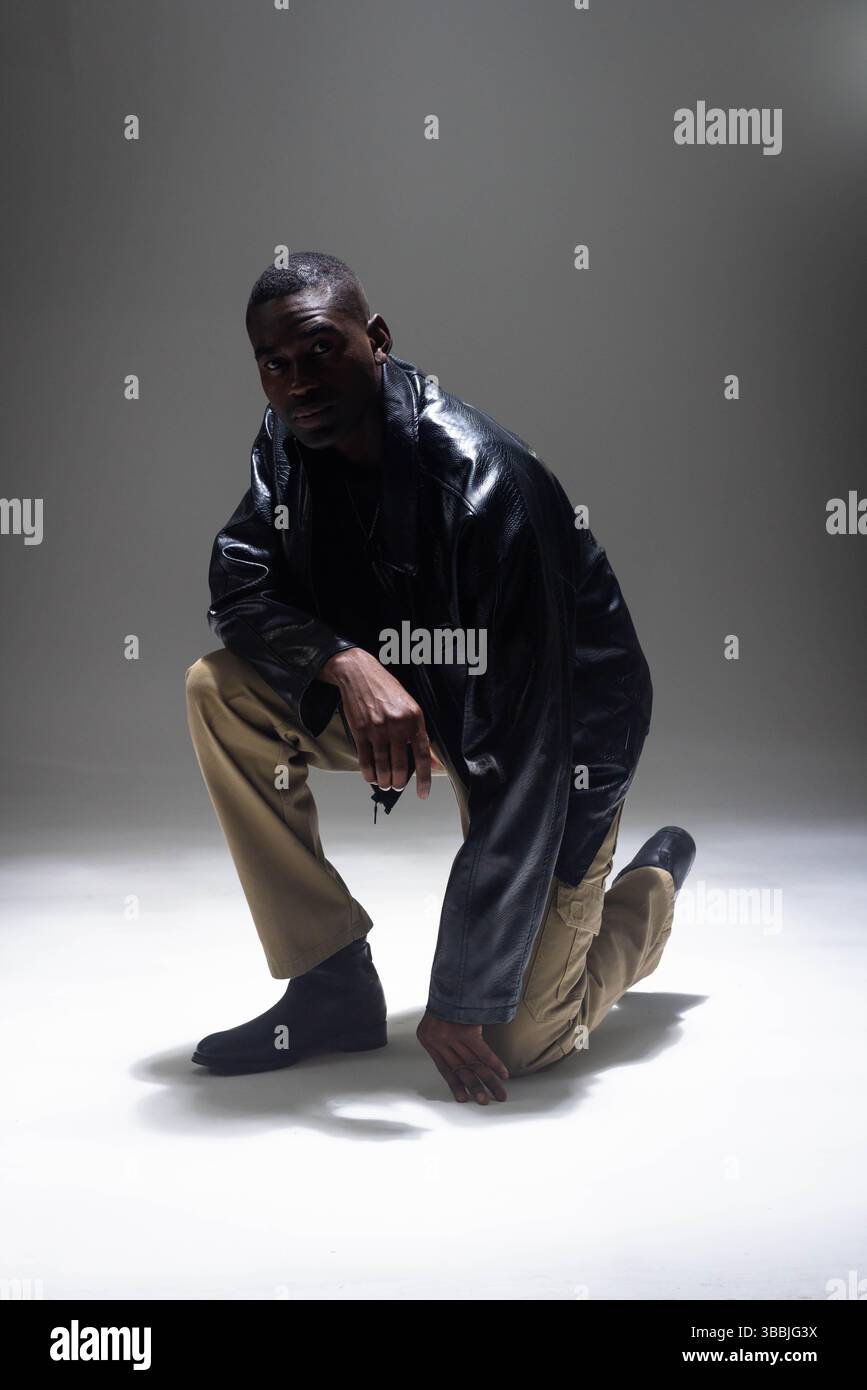 Uomo afro-americano inginocchiato in studio, con giacca in pelle nera, pantaloni cargo tan e stivali neri Foto Stock