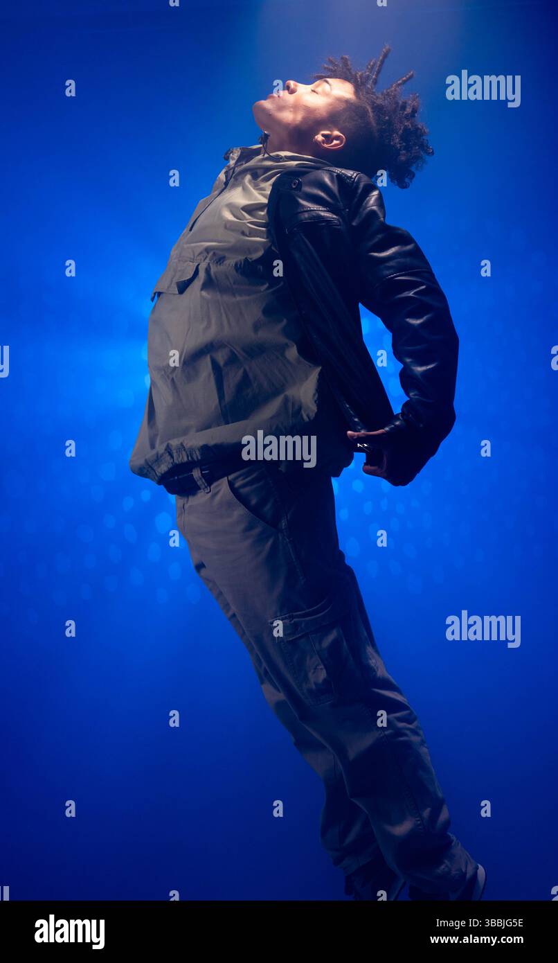 Esecutore maschile pendente che posa drammaticamente sul palco, con giacca con cappuccio, pantaloni cargo e foschia blu Foto Stock Esecutore maschile pendente che posa drammaticamente sul palco, con giacca con cappuccio, pantaloni cargo e foschia blu Foto Stock