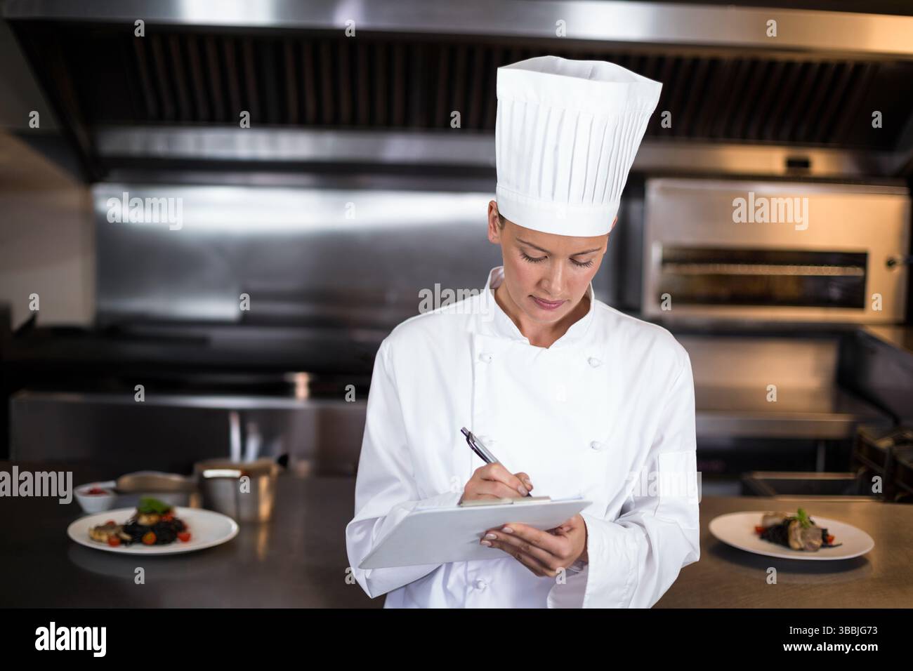 Scrivendo una chef donna annotando i dettagli sugli appunti nella cucina commerciale, con piatti guarniti Foto Stock