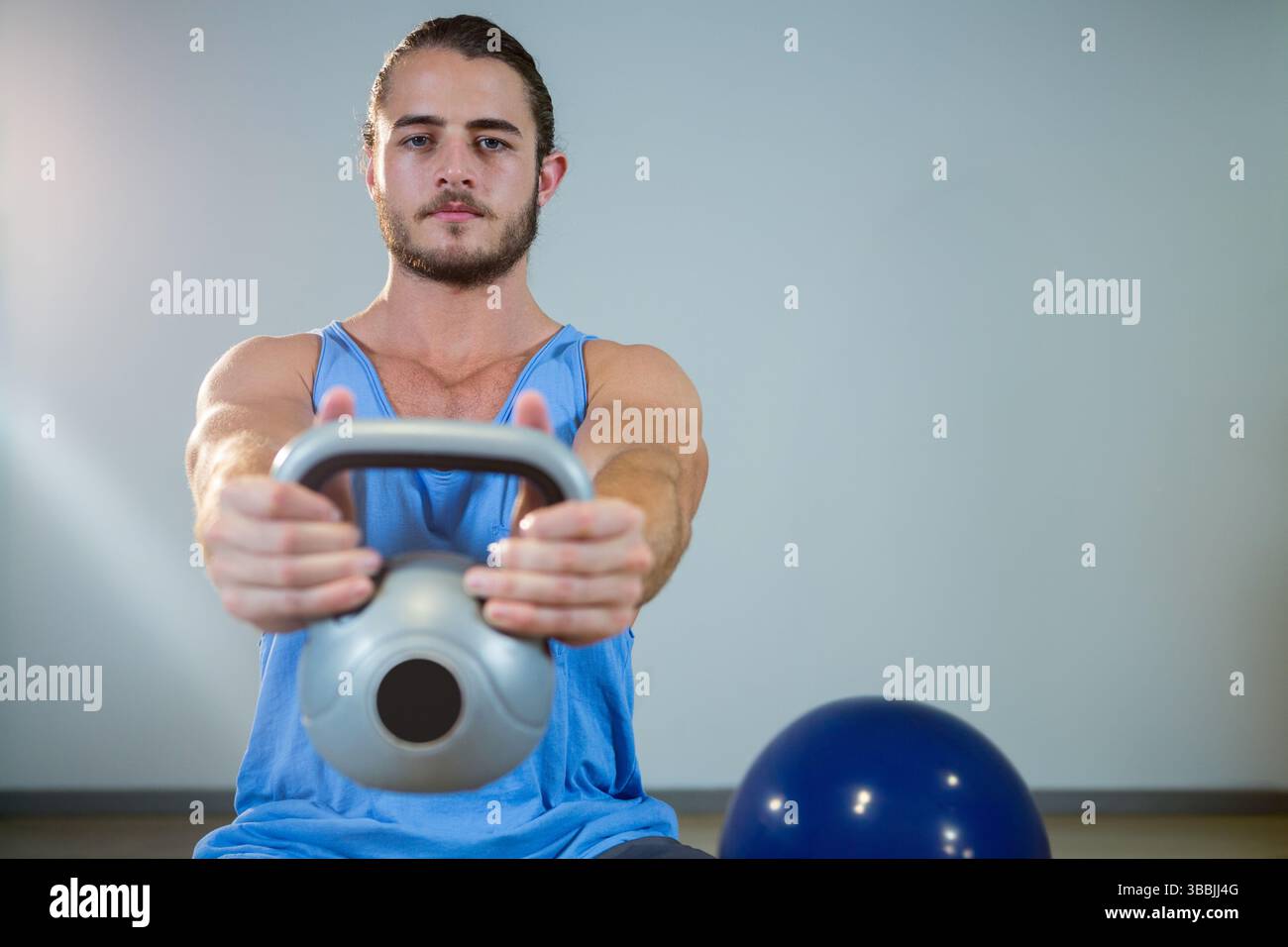 Uomo che solleva il kettlebell all'altezza del petto in palestra, indossa una canotta blu con palla da ginnastica Foto Stock