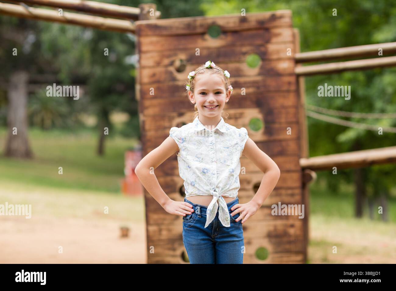 Bambina sorridente in piedi con le mani sui fianchi nel parco all'aperto, con parete da arrampicata e fascia floreale Foto Stock