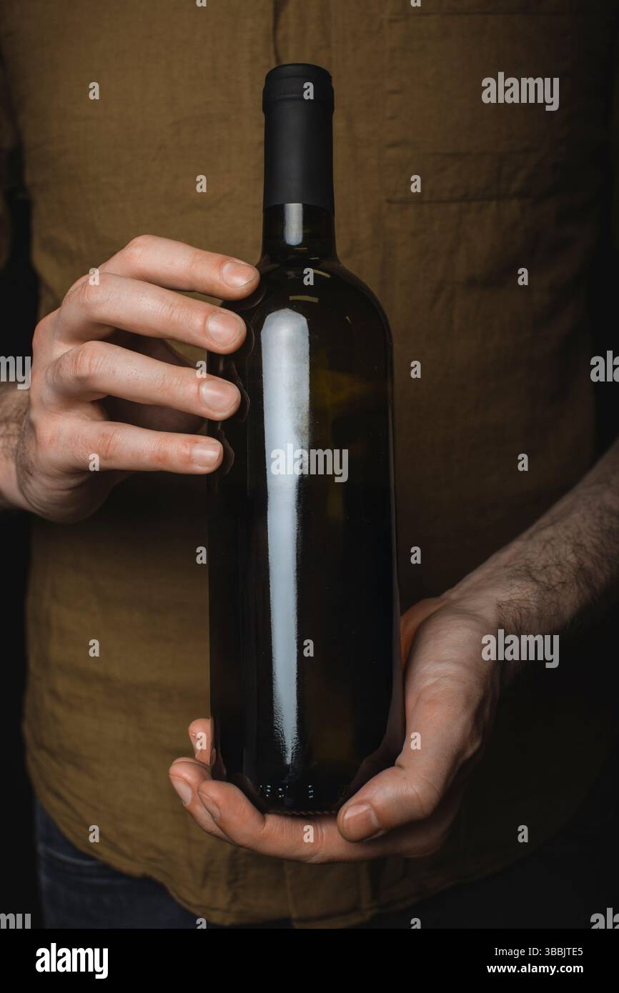 Bottiglia di vino senza etichetta tenuta per mano su uno sfondo scuro della camicia. Perfetto per i mockup di marca del vino con un'atmosfera autentica e moderna. Foto Stock