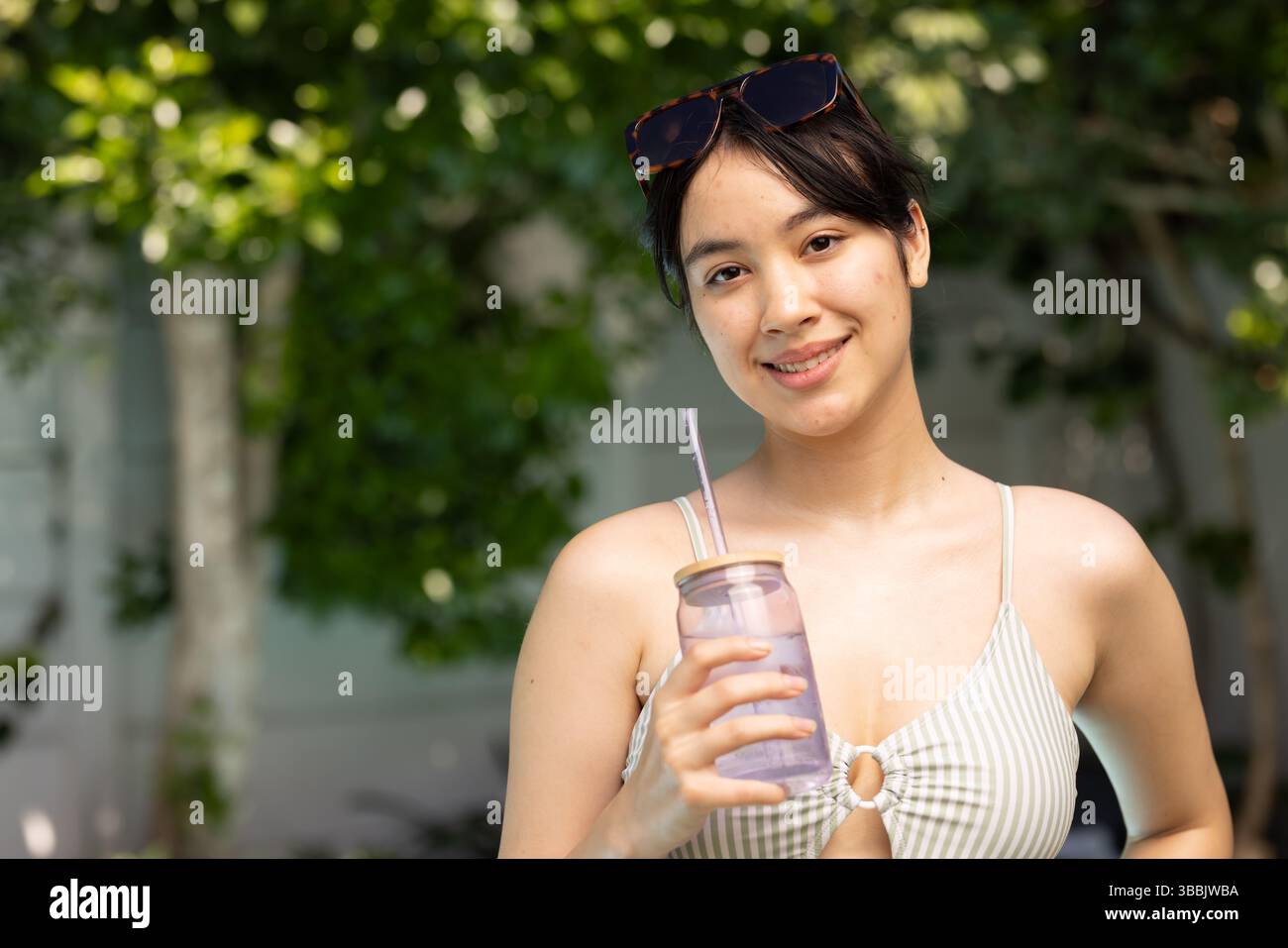 Sorseggiando una donna asiatica che indossa un bikini a righe in giardino, con bicchiere trasparente e occhiali da sole Foto Stock