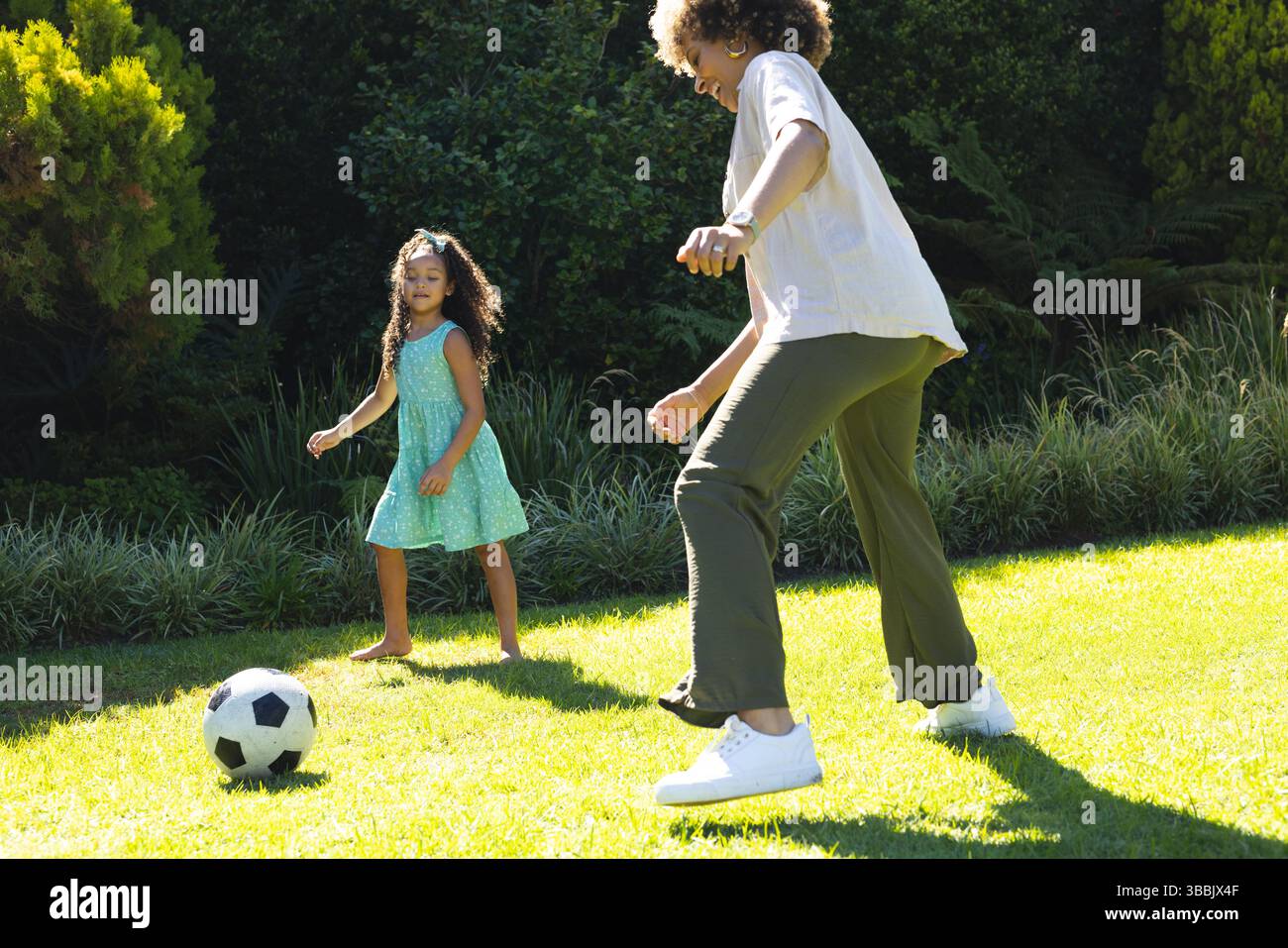Madre e figlia afroamericane giocano a calcio su un prato verde illuminato dal sole, con pallone da calcio Foto Stock