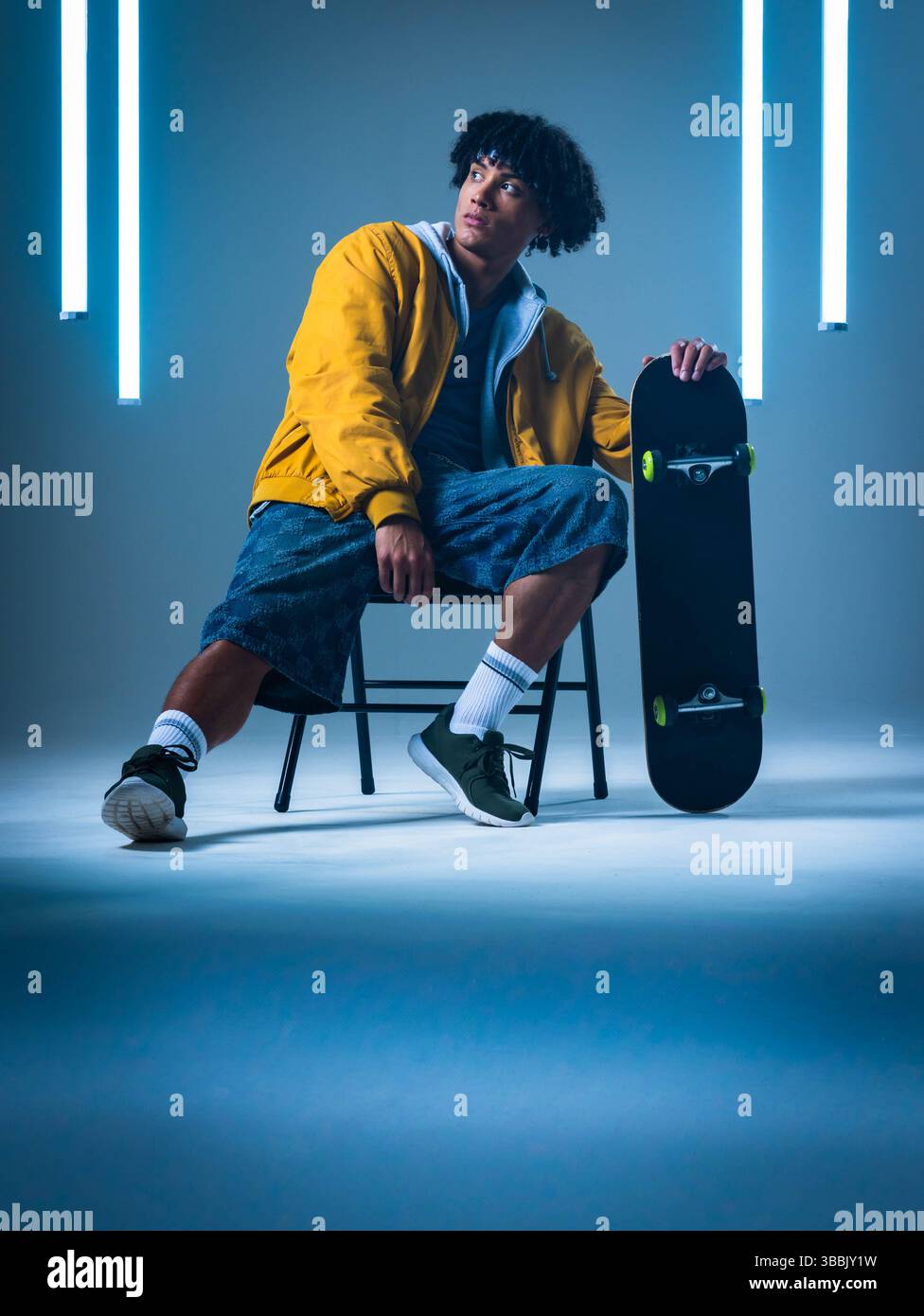Uomo afro-americano seduto che indossa un bomber in studio, regge lo skateboard con luci al neon Foto Stock