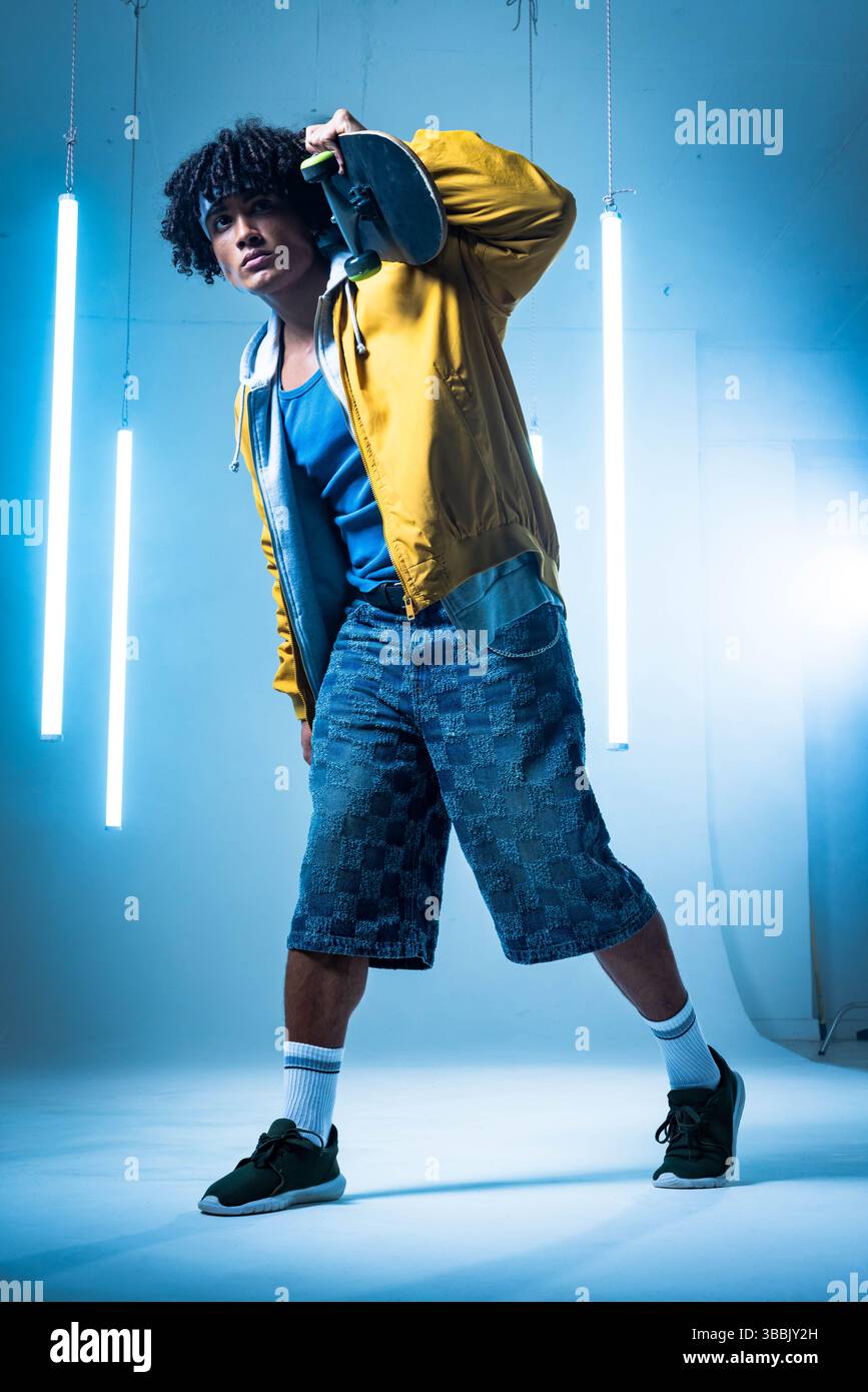 Posa di un uomo afroamericano che tiene lo skateboard dietro il collo in uno studio fotografico con tubi al neon Foto Stock