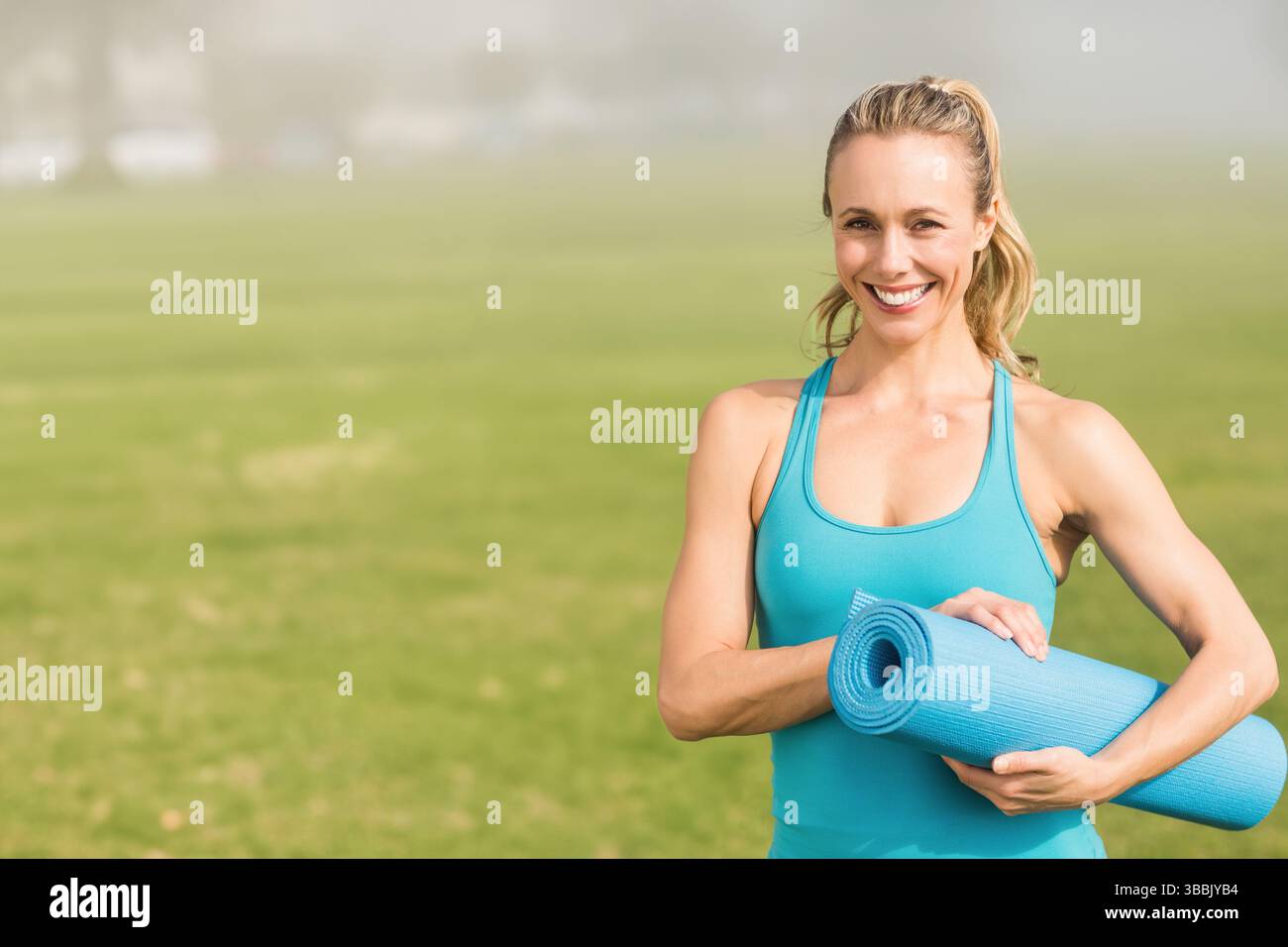 Donna sorridente che tiene in mano un tappetino da yoga blu arrotolato nel nebbioso parco erboso di mattina presto, spazio fotocopie Foto Stock