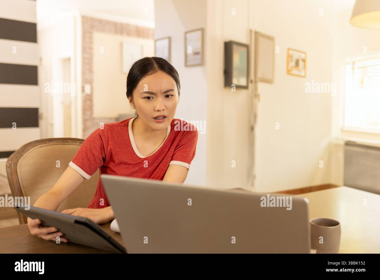 Giovane donna asiatica appoggiata a un tavolo di legno in casa, che utilizza un computer portatile e un tablet con tazza da caffè Foto Stock