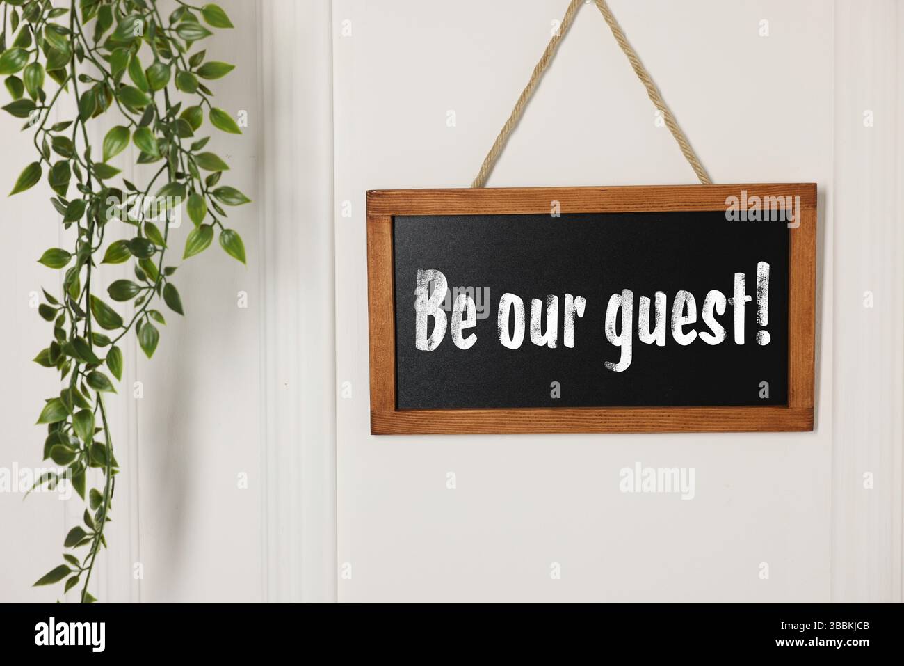 Piccola lavagna con la frase "Be Our Guest" e la pianta della casa appesa su una parete bianca Foto Stock