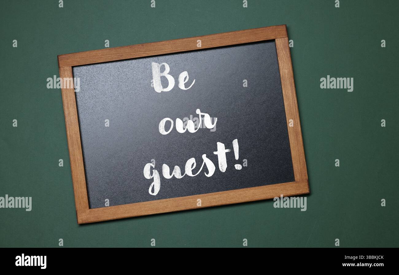 Piccola lavagna con frase Be Our Guest su sfondo verde scuro, vista dall'alto Foto Stock