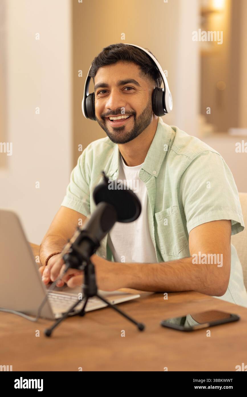 Giovane uomo indiano adulto che registra in studio domestico, scrivendo sul computer portatile con cuffie e microfono Foto Stock