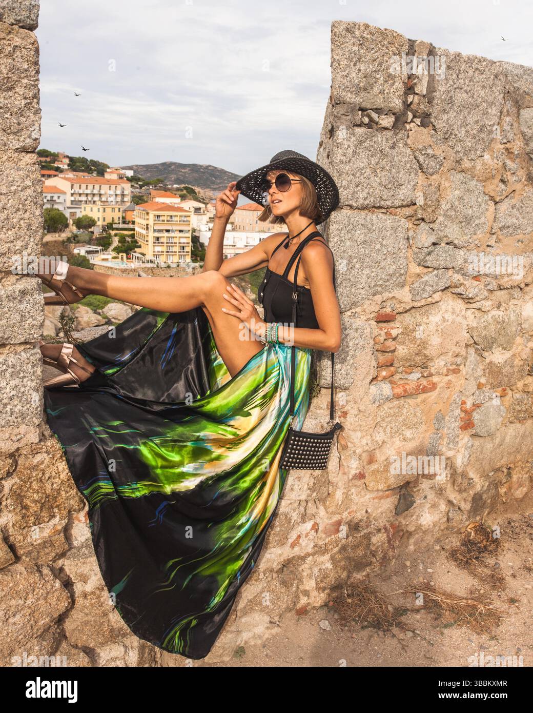 La donna alla moda è seduta sul muro di pietra della splendida città storica di Calvi, nell'isola della Corsica, in Francia. Indossa outfi eleganti e femminili. Foto Stock