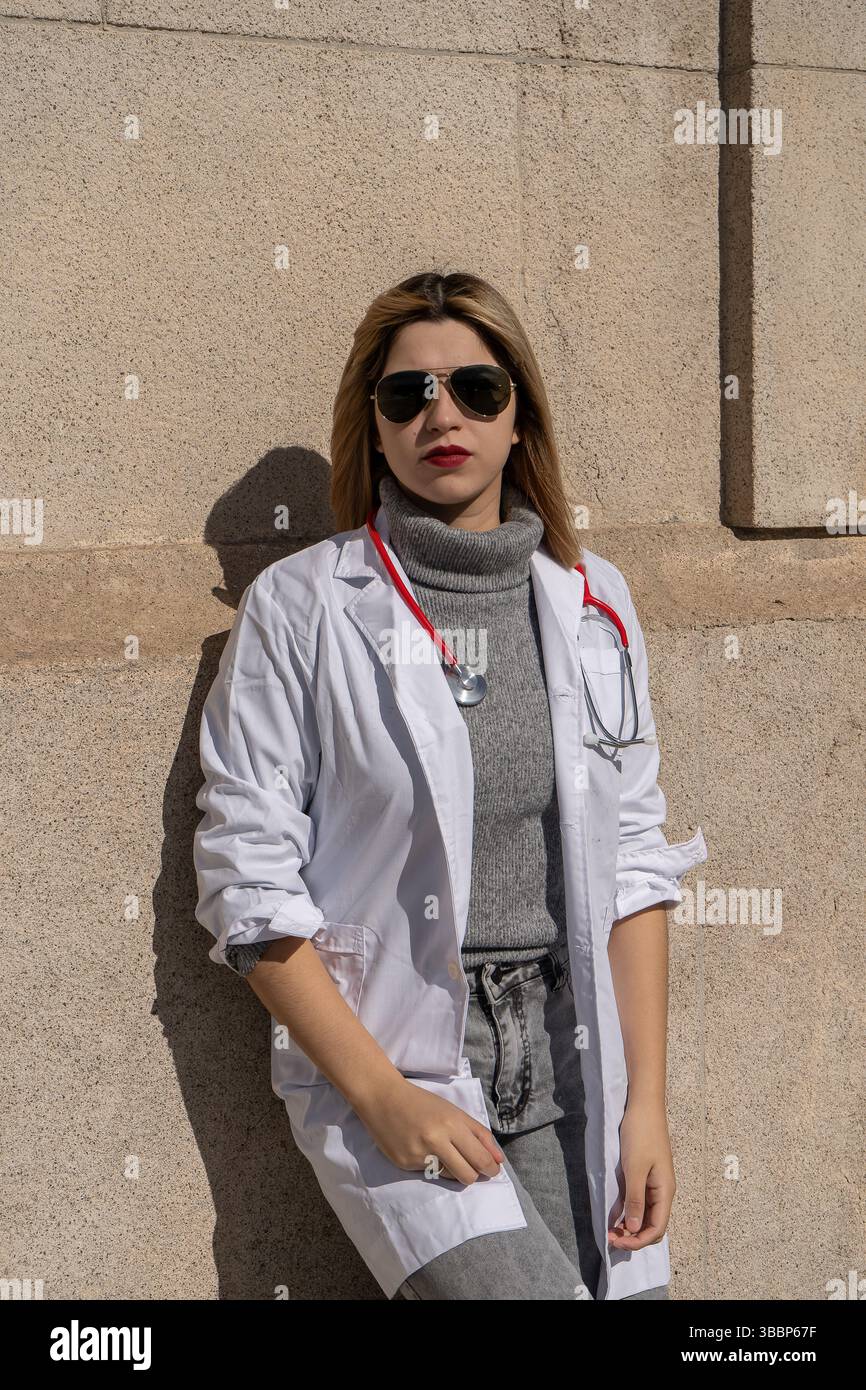 Giovane e sicura medico donna con stetoscopio e occhiali da sole che posano contro una parete Foto Stock